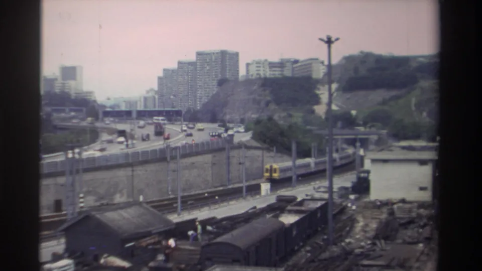 Aged Super 8 footage from Taiwan, 1982: wide view of railway, highway traffic, concrete walls, hillside apartments, and freight sheds with characteristic pink-hued film fade.