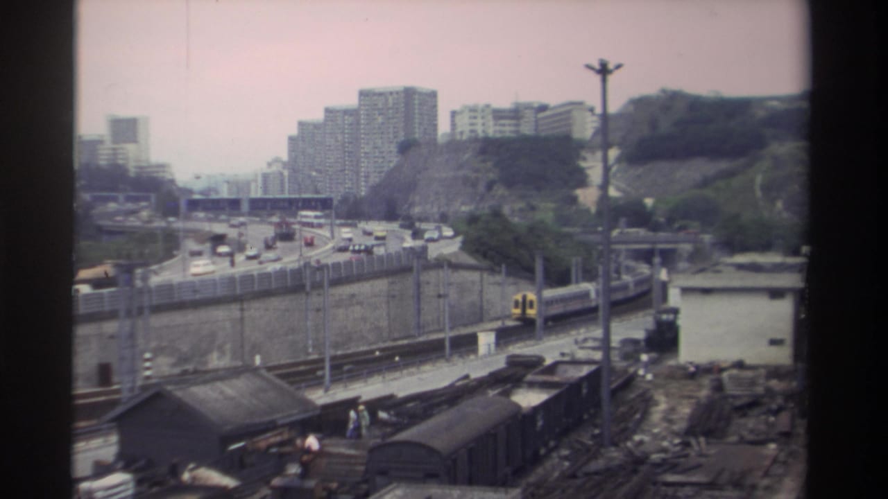 Yellow and white passenger train passing a marshalling yard beside a busy highway, Taiwan, circa 1982; dense high-rise blocks visible on tree-covered hillside.