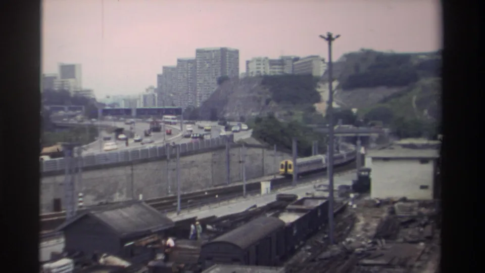 Yellow and white passenger train passing a marshalling yard beside a busy highway, Taiwan, circa 1982; dense high-rise blocks visible on tree-covered hillside.