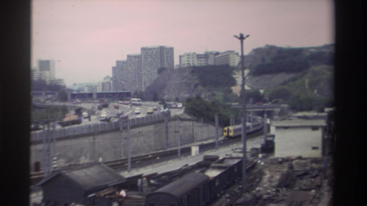 Passenger train moving along tracks parallel to a busy highway, freight wagons in foreground, high-rise buildings on distant hillside.