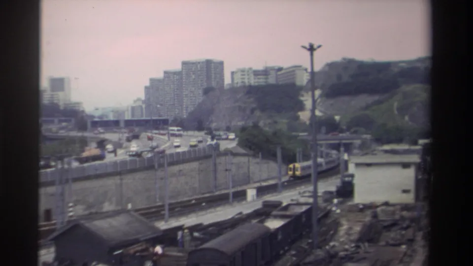 8mm frame showing a yellow and white train beside a highway with heavy traffic, Taiwan, circa 1982; residential blocks visible on the hillside behind.