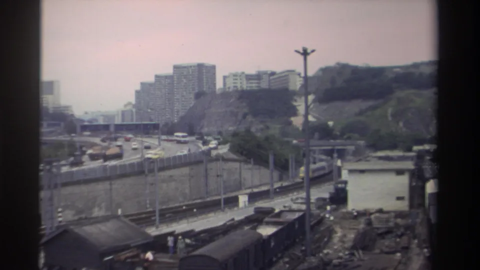 8mm frame of a Taiwanese railway yard in 1982 — freight cars and a yard shed in the foreground, busy highway in mid-ground, residential tower blocks on the hillside.