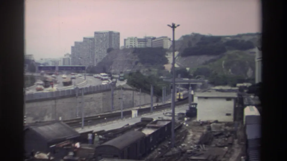 Late frame from an 8mm travel shot in Taiwan, circa 1982, showing freight wagons and a yard building in the foreground, cars on a multi-lane highway, and residential high-rises on a vegetated hillside.