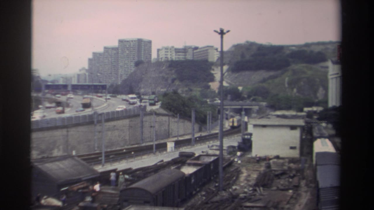 8mm home movie frame, c.1982 Taiwan: freight wagons in foreground, curved multi-lane motorway with period cars, and concrete residential towers on a hillside.