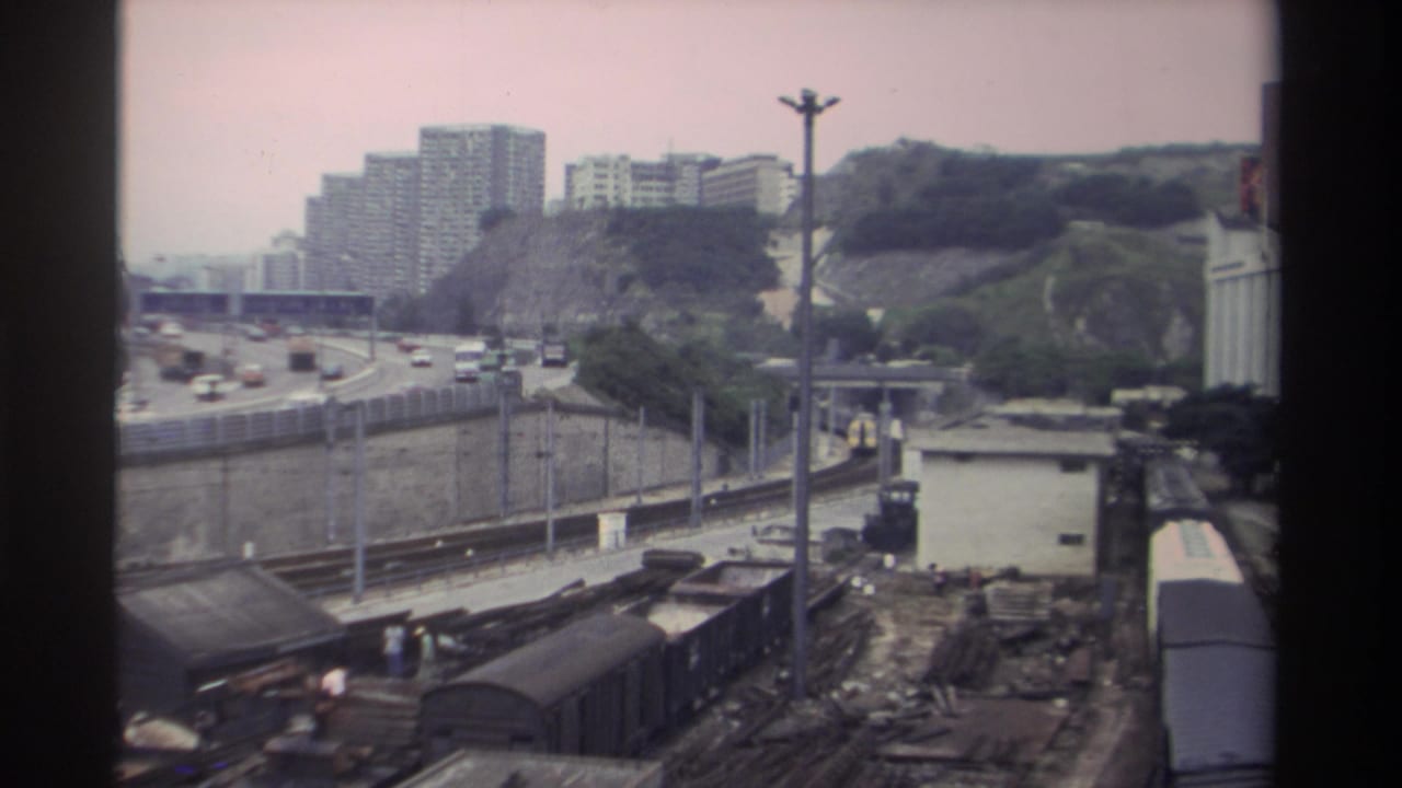 Similar elevated wide shot of the freight rail yard, motorway and hillside residential towers; composition nearly identical to preceding frame.