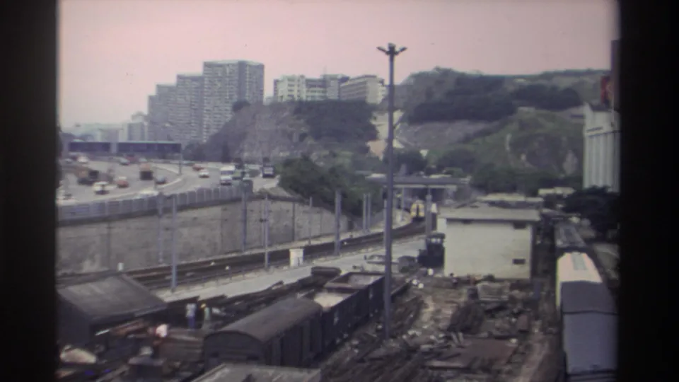 8mm frame, c.1982 Taiwan: freight wagons, curved motorway with period vehicles, concrete apartment towers on hillside, small figures visible near train cars.