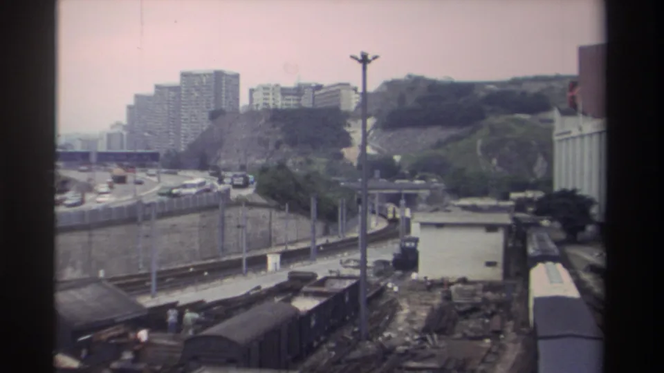 8mm frame, c.1982 Taiwan: white passenger rail car entering right of frame alongside freight wagons, motorway traffic and hillside residential towers behind.