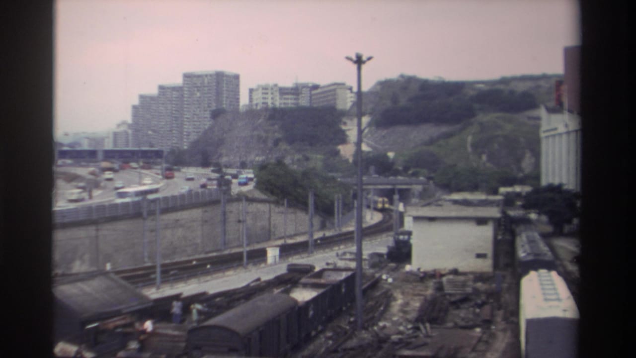 Super 8 footage from Taiwan, circa 1982, showing freight rail cars in a yard below a curved elevated highway, with apartment blocks on a hazy hillside beyond.
