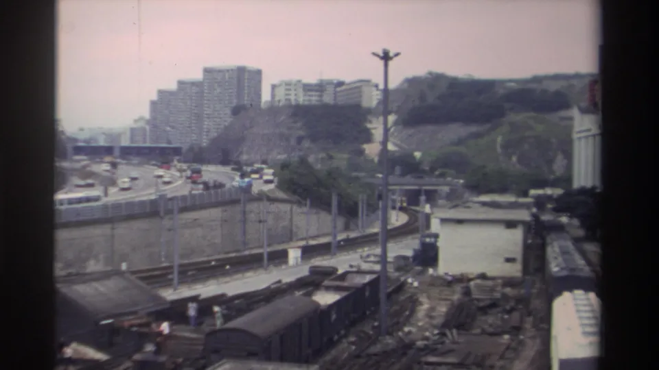 8mm film frame from Taiwan, circa 1982, capturing a rail yard, a curving elevated expressway with period vehicles, and densely built hillside housing estates.