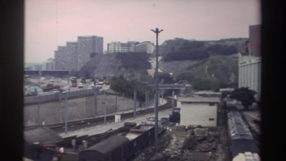 Final 8mm frame from Taiwan, circa 1982, showing freight cars, a raised urban expressway, densely packed hillside housing, and a red-orange structure visible at the right edge.