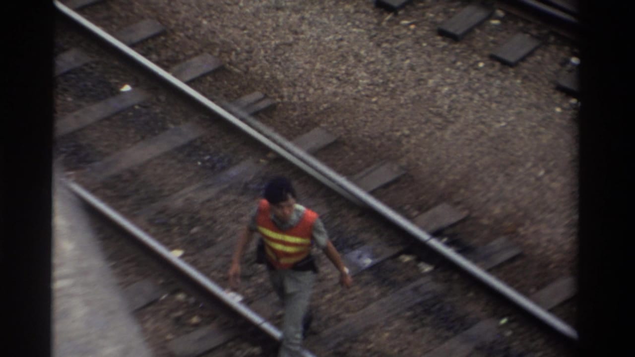 A railway maintenance worker in an orange and yellow high-visibility vest walks between steel rails in Taiwan, 1982, shot from an elevated angle on 8mm film.