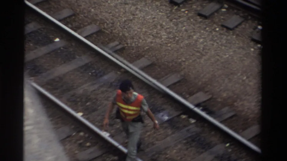 A railway maintenance worker in an orange and yellow high-visibility vest walks between steel rails in Taiwan, 1982, shot from an elevated angle on 8mm film.