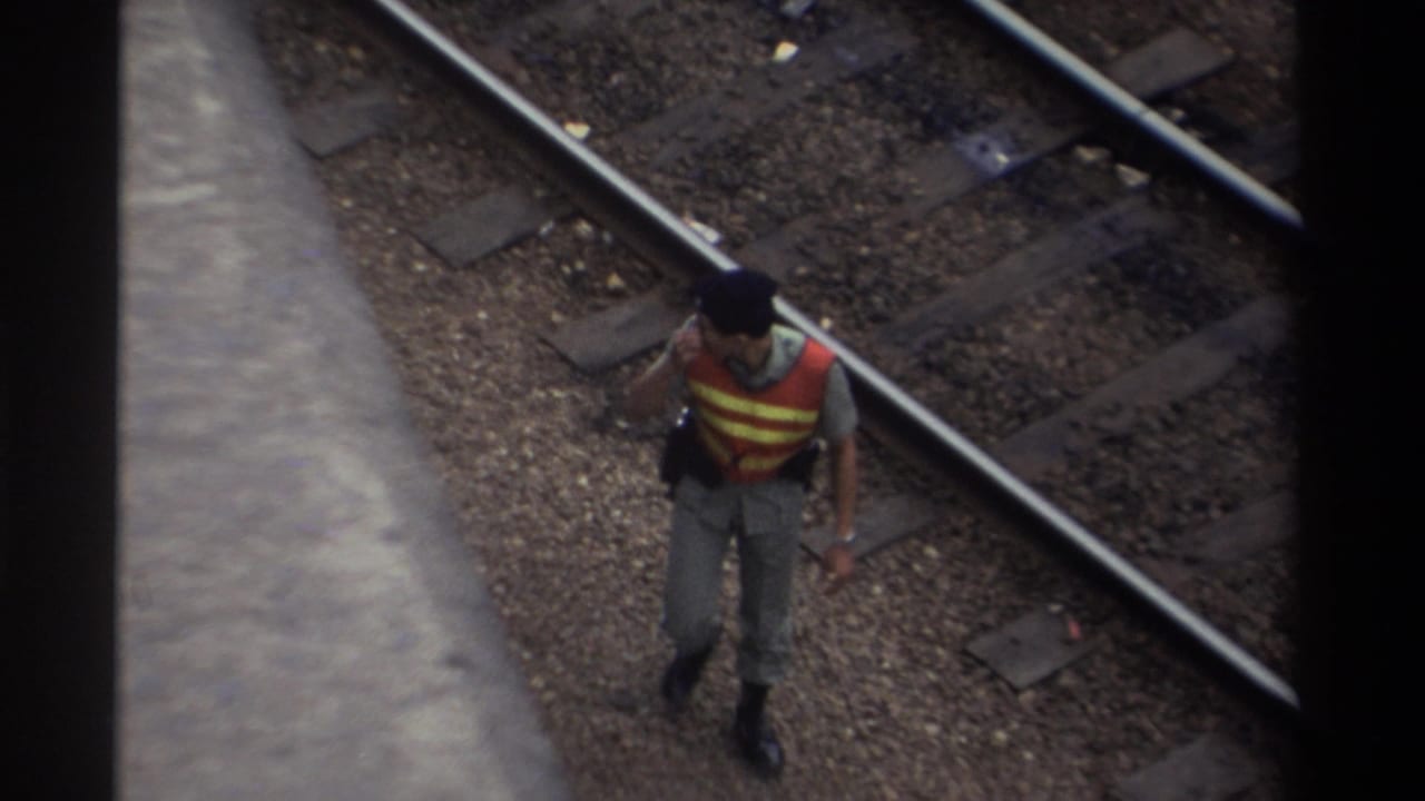 A railway worker in an orange high-visibility vest pauses mid-step with hand near face, viewed from above on a multi-track line, Taiwan 1982, 8mm film.