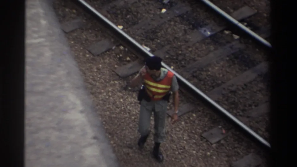 A railway worker in an orange high-visibility vest pauses mid-step with hand near face, viewed from above on a multi-track line, Taiwan 1982, 8mm film.