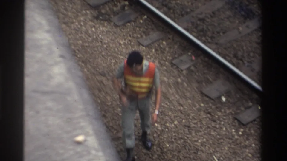The same railway worker walks forward along the ballast between rails, orange vest prominent against grey gravel, Taiwan 1982, 8mm film, overhead shot.