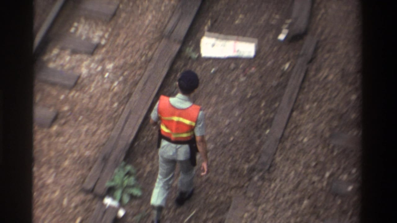 High-angle view of a man in an orange safety vest walking away along railway tracks laid on gravel ballast.