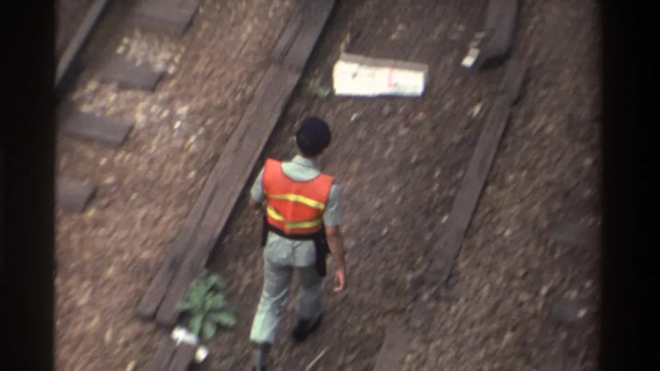 Railway worker in orange reflective safety vest and grey uniform walks between parallel tracks in Taiwan, filmed from above on 8mm in 1982.