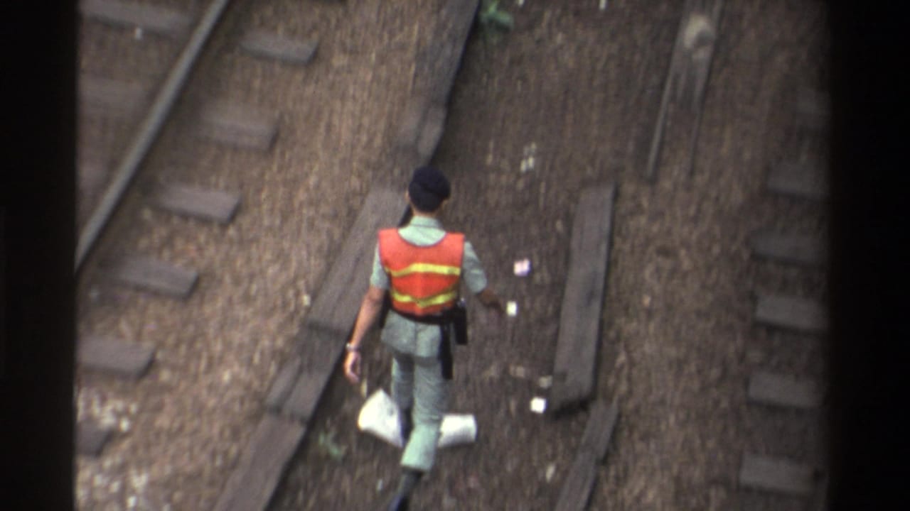 High-angle view of a railway worker in orange safety vest walking between tracks, a white bag visible at his side.