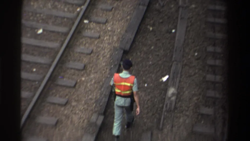 Railway worker in orange reflective vest walks between parallel tracks on gravel ballast, 8mm film from Taiwan, 1982.