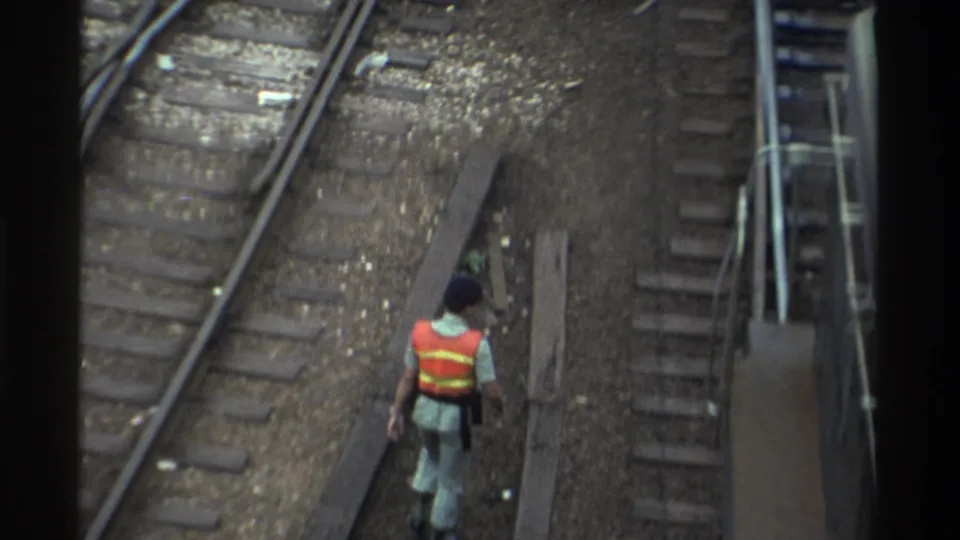 Railway worker in orange-and-yellow safety vest crossing parallel steel tracks in a rail yard, Taiwan, circa 1982; 8mm footage shot from an elevated position.