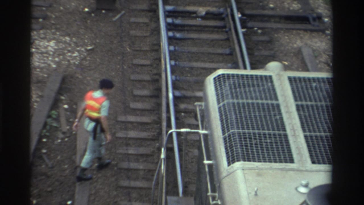 Rail worker in orange-and-yellow vest moving along tracks adjacent to a concrete access structure with metal grating, Taiwan, circa 1982; 8mm overhead footage.