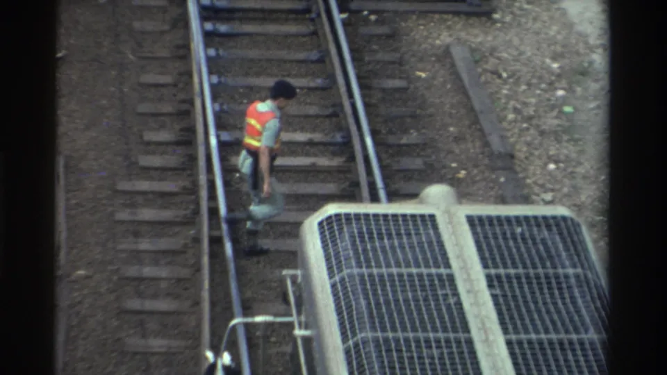 Rail worker in orange-and-yellow safety vest walking away between tracks with a concrete structure featuring metal grating in the foreground, Taiwan, circa 1982.