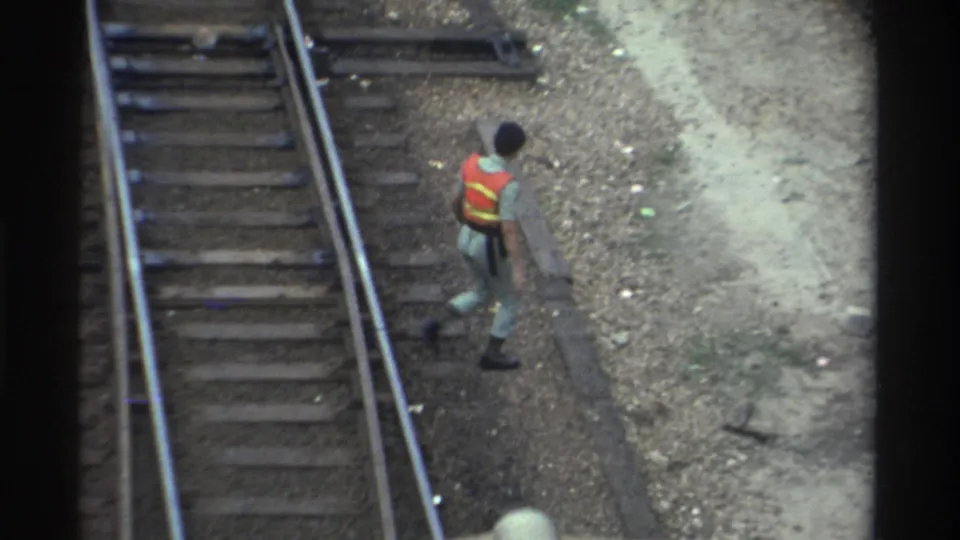 Rail maintenance worker in orange-and-yellow vest walking along tracks with a concrete structure to the left, Taiwan, circa 1982; 8mm overhead perspective.