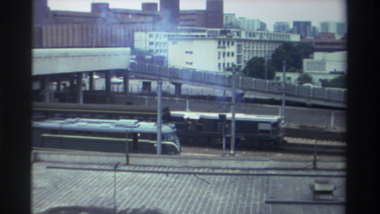 8mm home movie frame, Taiwan c. 1982 — diesel train traveling past an elevated highway with white residential buildings and trees in the background.