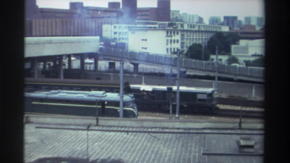 8mm home movie frame, Taiwan c. 1982 — diesel train traveling past an elevated highway with white residential buildings and trees in the background.