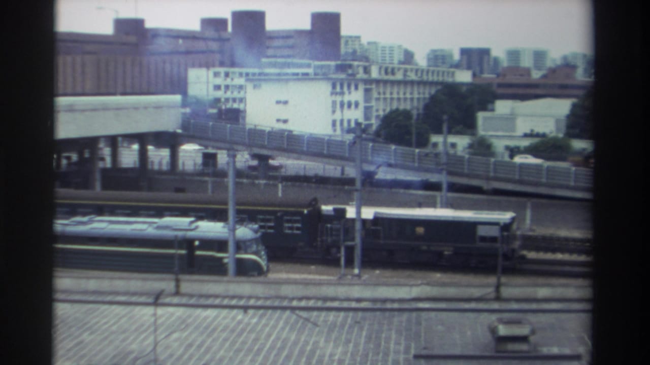 8mm frame, Taiwan c. 1982 — locomotive advances along rail corridor with elevated road structure and dense white urban buildings visible in background.