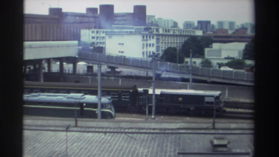 8mm frame, Taiwan c. 1982 — locomotive advances along rail corridor with elevated road structure and dense white urban buildings visible in background.