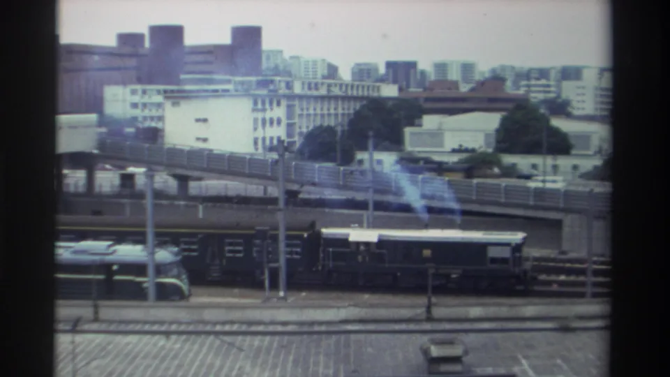 8mm home movie frame, Taiwan c. 1982 — train cars below elevated highway with high-rise blocks and trees behind; blue haze from exhaust or film artifact.