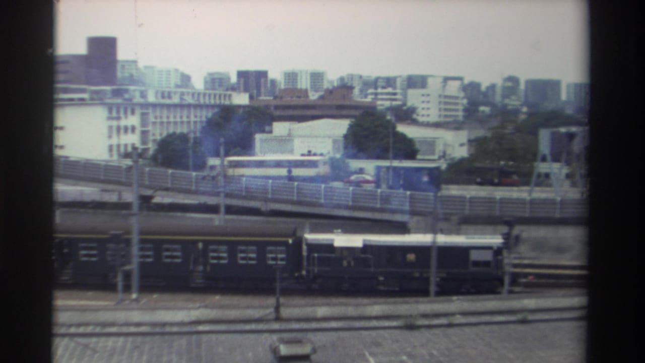 Elevated view over a Taiwanese rail yard circa 1982, showing dark train carriages, a curved overpass with a yellow vehicle, and a mid-rise cityscape under overcast skies.