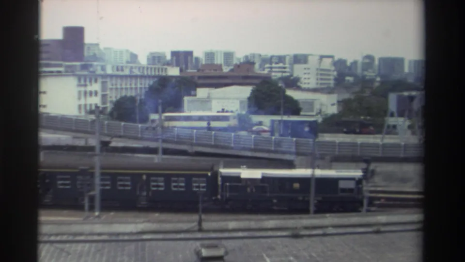 Elevated view over a Taiwanese rail yard circa 1982, showing dark train carriages, a curved overpass with a yellow vehicle, and a mid-rise cityscape under overcast skies.