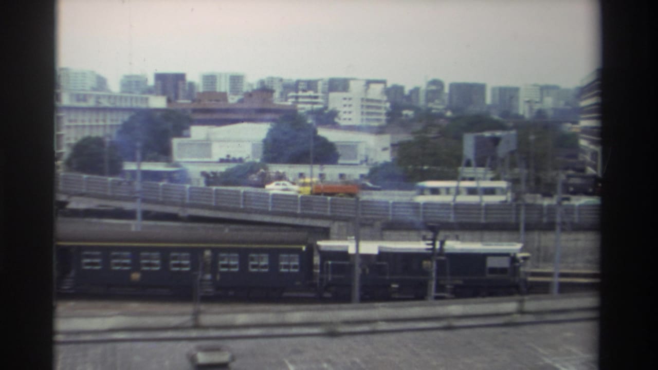 Elevated view over the same Taiwanese rail yard, circa 1982, with a yellow-orange vehicle moving on the overpass above dark train carriages and a mid-rise skyline.