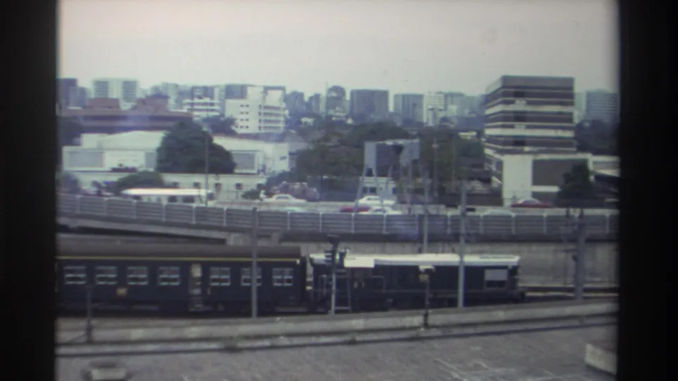 Wider elevated shot of the same Taiwanese urban transport scene, circa 1982, with red vehicles on the elevated road, a pylon structure mid-ground, and a dense mid-rise skyline.