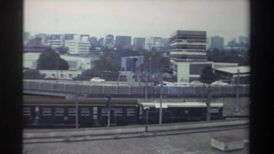 Final frame of the Taiwanese rail-and-highway scene, circa 1982, showing mid-rise commercial and residential blocks above dark train carriages, elevated roadway, and trees.