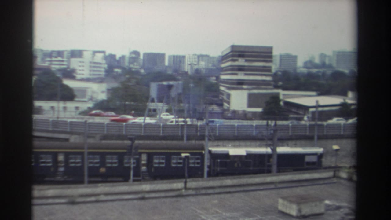 Dark passenger train on ground-level tracks passing below an elevated road overpass; mid-rise commercial buildings and high-rise towers in background, Taiwan 1982.