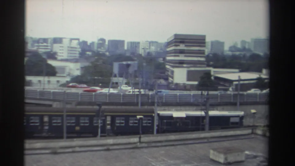 Dark passenger train on ground-level tracks passing below an elevated road overpass; mid-rise commercial buildings and high-rise towers in background, Taiwan 1982.