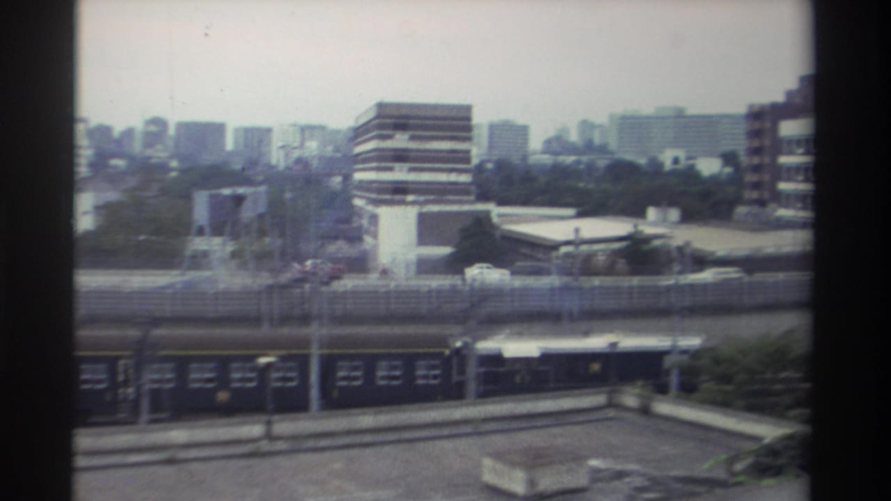 Continuation shot of the dark passenger train beneath an elevated roadway; urban commercial and residential skyline stretching across the background, Taiwan 1982.