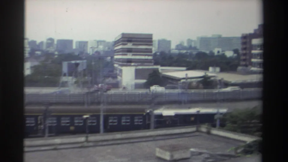 Continuation shot of the dark passenger train beneath an elevated roadway; urban commercial and residential skyline stretching across the background, Taiwan 1982.