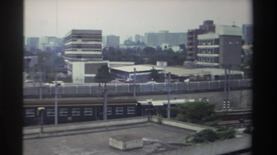 Wide elevated view of a Taiwanese urban district after the train has passed; commercial blocks, scattered trees, and residential towers visible in background, Taiwan 1982.