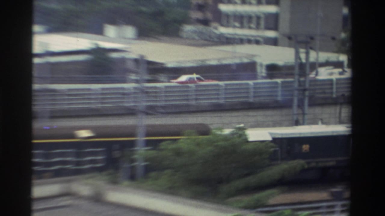 Dark green passenger train with yellow stripe moves through an urban rail corridor in Taiwan, circa 1982, shot on 8mm from an elevated window.