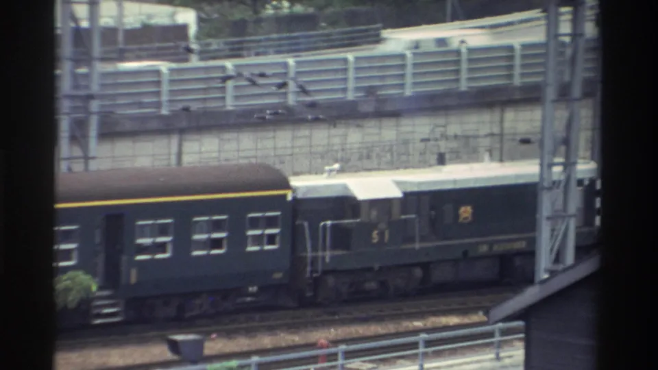 Dark green train numbered 51 moves through an urban corridor in Taiwan, circa 1982; blue road sign and small background figures visible.