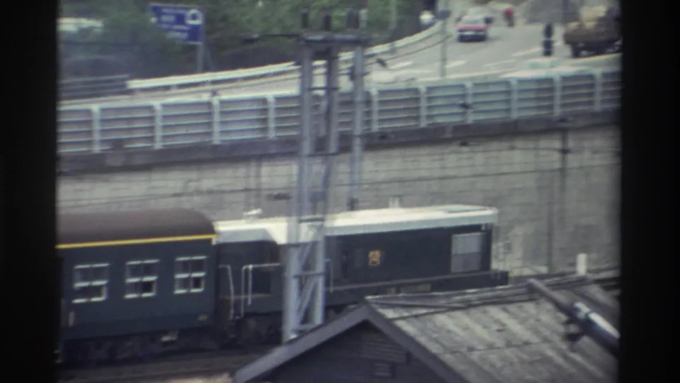 Dark green passenger cars with yellow stripe continue through an urban rail corridor in Taiwan, circa 1982; a rooftop structure is visible in the foreground.