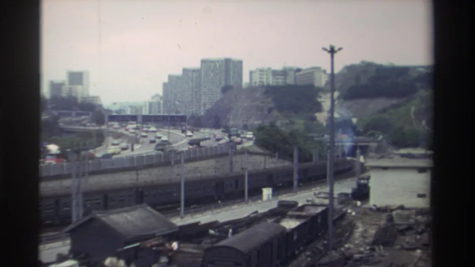 Wide view of a Taiwan transportation corridor, c. 1982 — train tracks, multi-lane expressway with traffic, and concrete apartment towers on a hillside, 8mm.