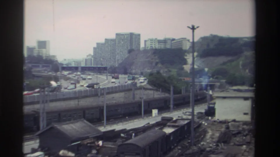 Elevated view of a Taiwanese expressway curving past a rail yard, low-rise storage sheds in the foreground and high-rise housing on the hillside, 8mm, c. 1982.