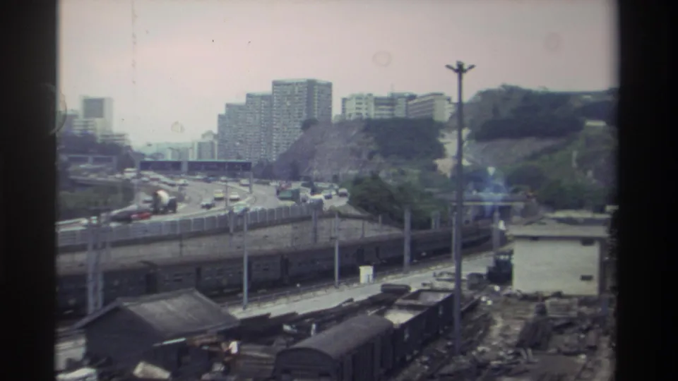 Continued elevated shot of a Taiwanese transportation corridor — expressway traffic, rail yard, and hillside housing blocks, 8mm home movie, c. 1982.