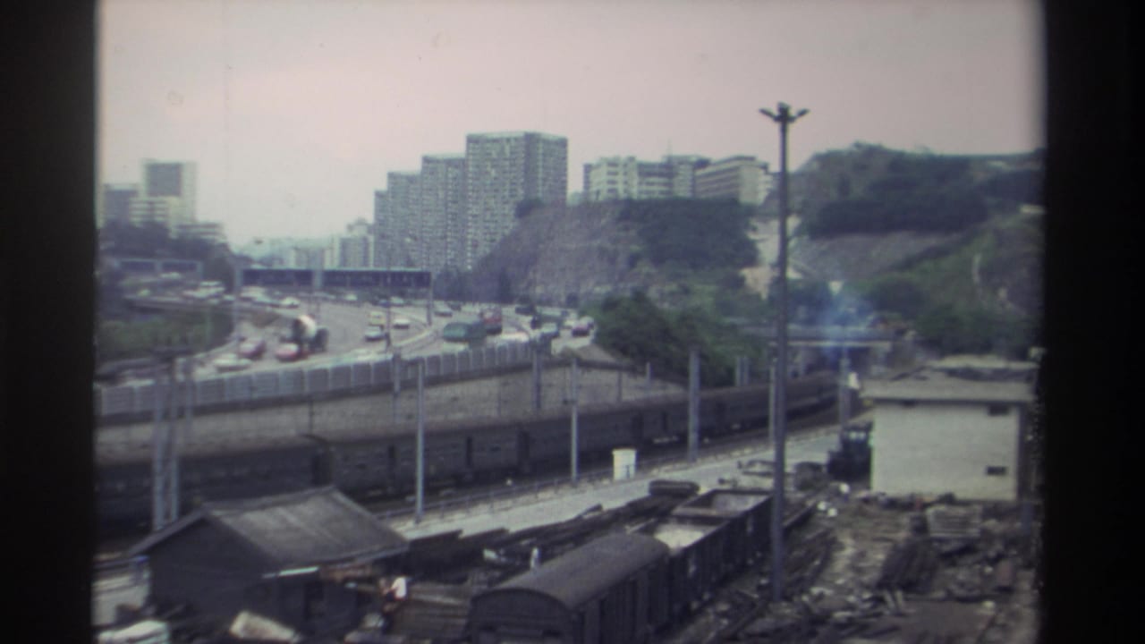 1982 Taiwan: 8mm window shot showing a rail freight yard, congested expressway, and residential high-rise towers on a green hillside under an overcast sky.