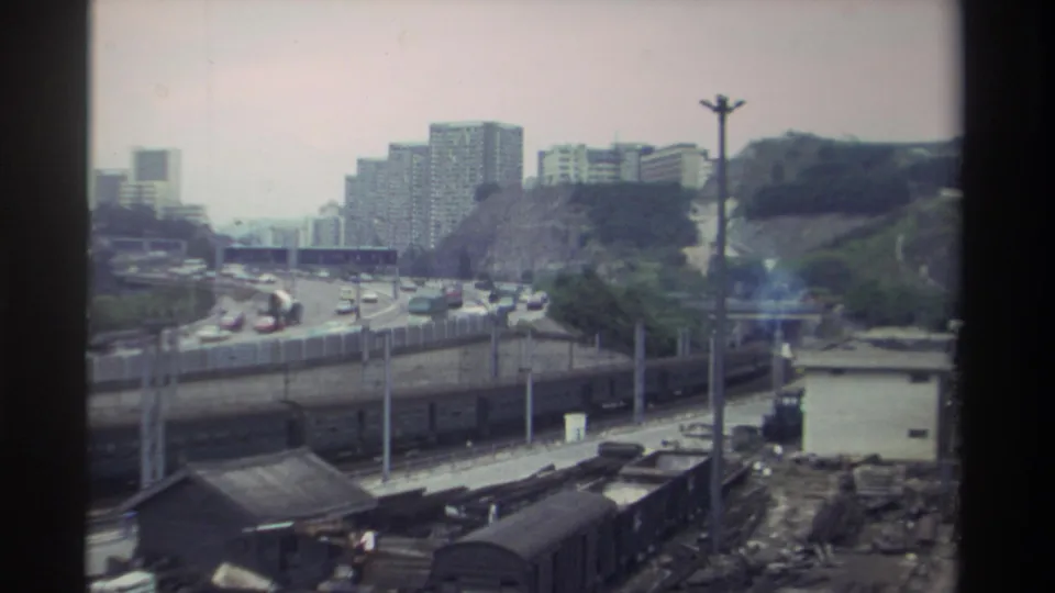 1982 Taiwan: 8mm window shot showing a rail freight yard, congested expressway, and residential high-rise towers on a green hillside under an overcast sky.